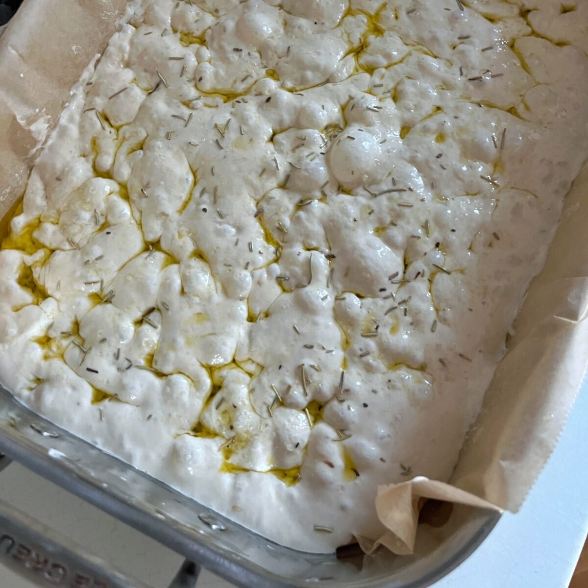 Unbaked focaccia dough in a stainless steel baking dish lined with parchment paper, drizzled with olive oil and sprinkled with sea salt.