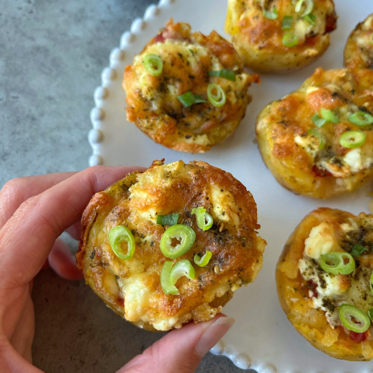 A hand holds a potato cup topped with cheese and sliced green onions—with several more cups arranged on a white plate in the background.