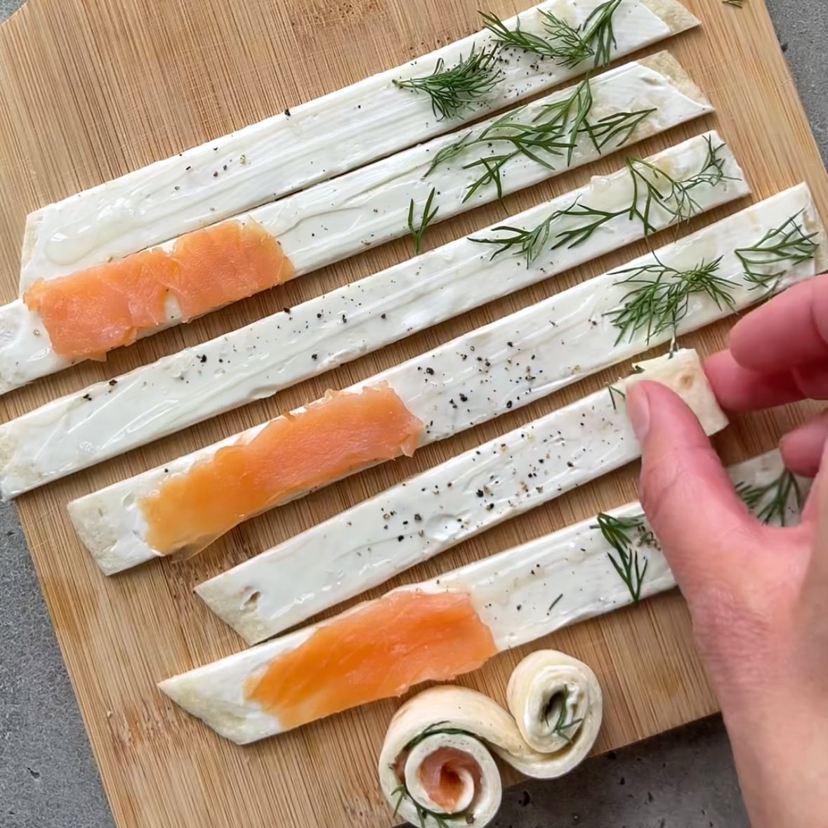 Tortilla strips topped with dill and half topped with smoked salmon are on a chopping board. A hand rolls up one of the strips.