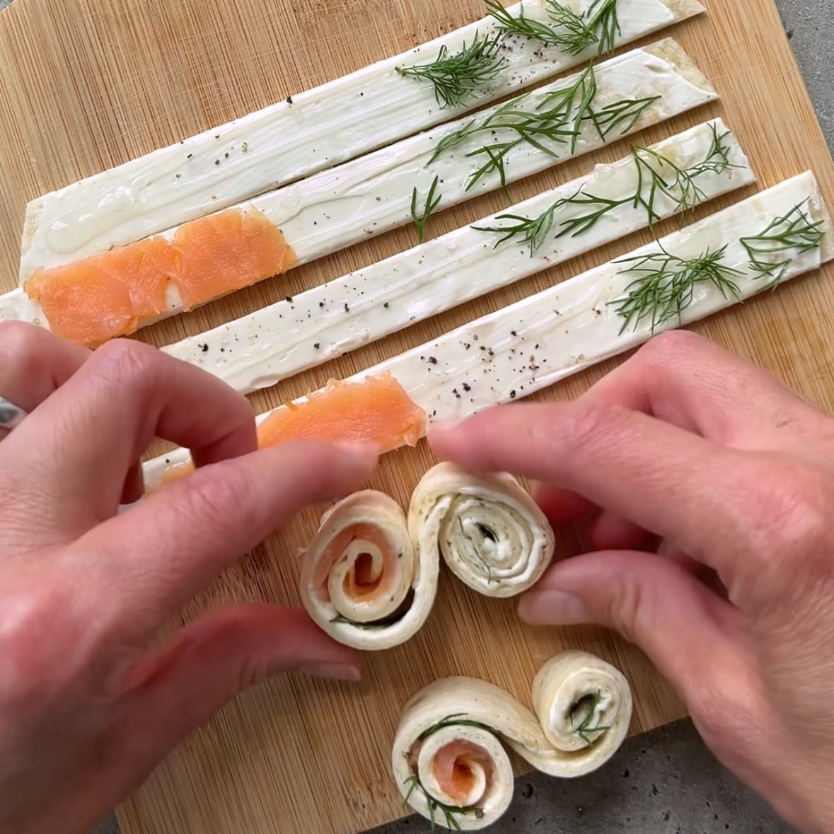 Two hands connect rolled up strips of tortilla topped with dill and smoked salmon to make elegant appetizers.