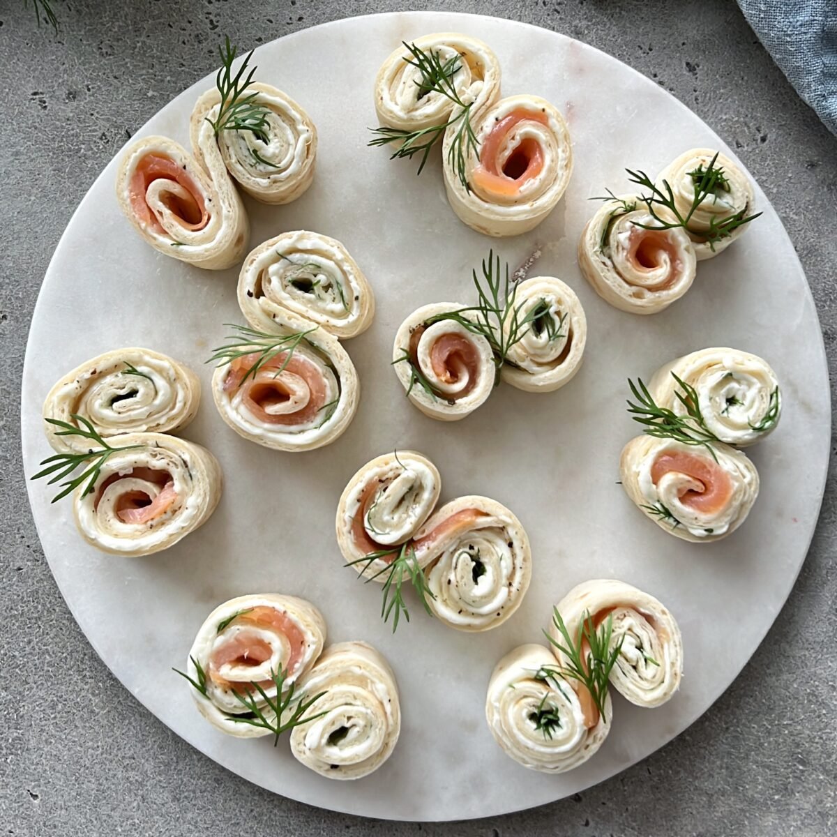 A marble plate with smoked salmon and cream cheese pinwheels, garnished with fresh dill, arranged in a circular pattern.
