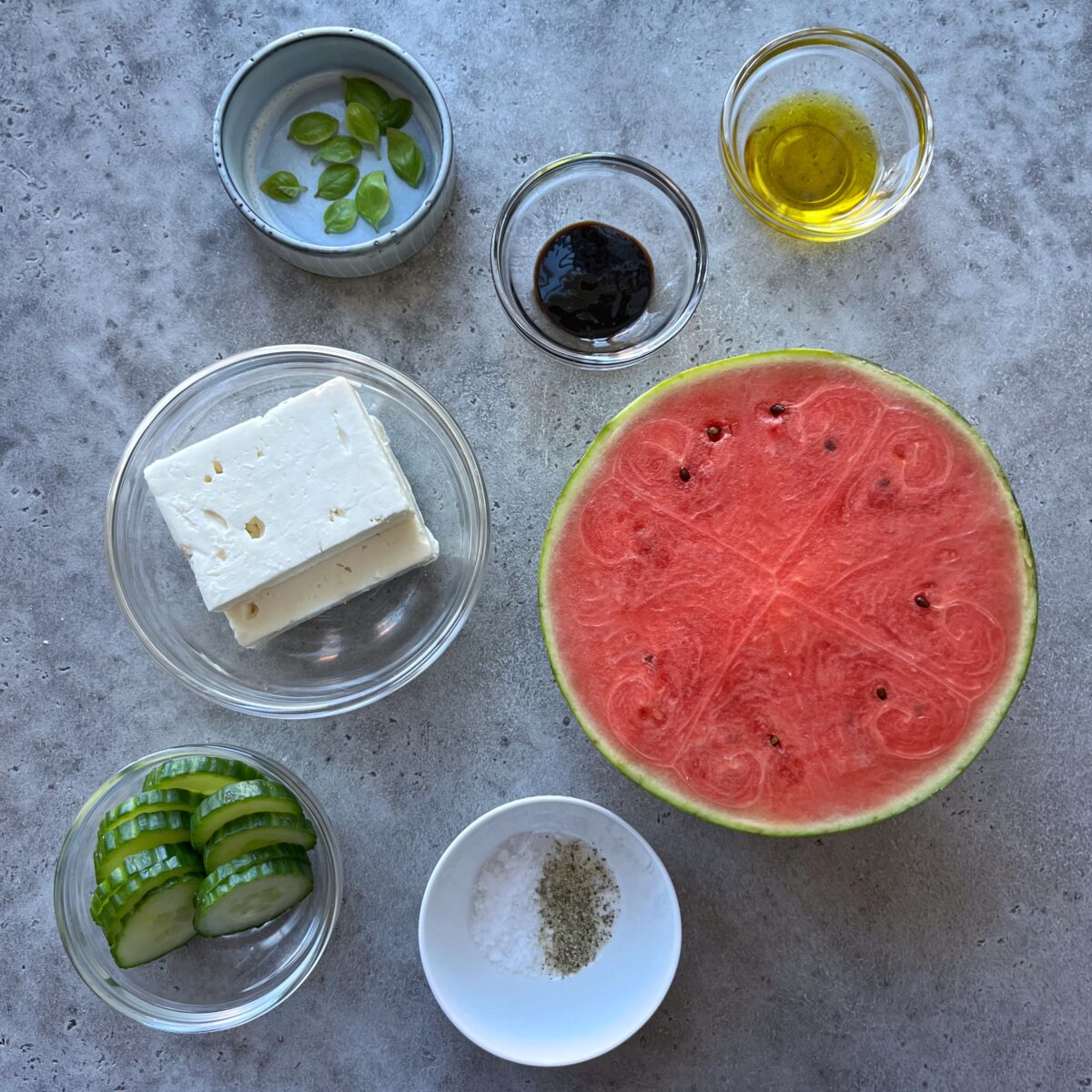 Ingredients for a Watermelon Cucumber Salad arranged on a gray surface: sliced cucumber, feta cheese, watermelon half, fresh basil, balsamic glaze, olive oil, salt, and pepper.