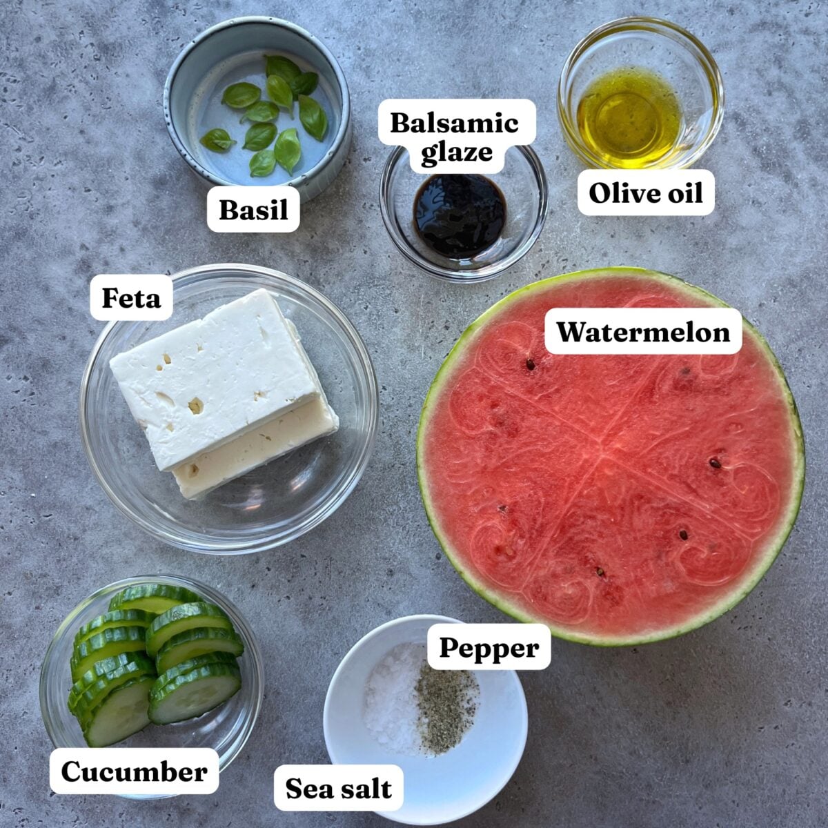 Ingredients for a Watermelon Cucumber Salad arranged on a gray surface featuring sliced cucumber, feta cheese, watermelon half, fresh basil, balsamic glaze, olive oil, salt, and pepper.