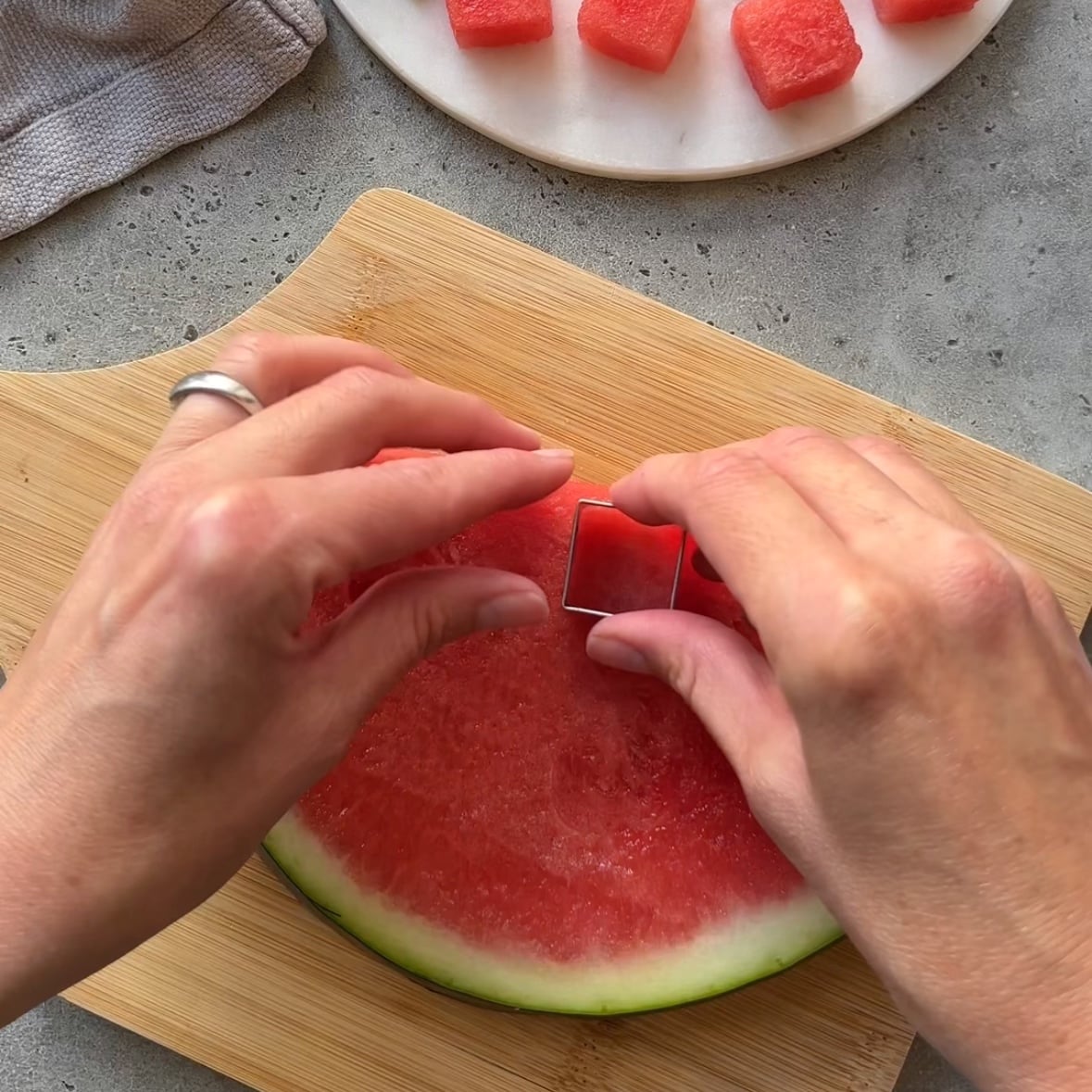 Two hands cut out a cube from a halved piece of watermelon with a square cookie cutter.