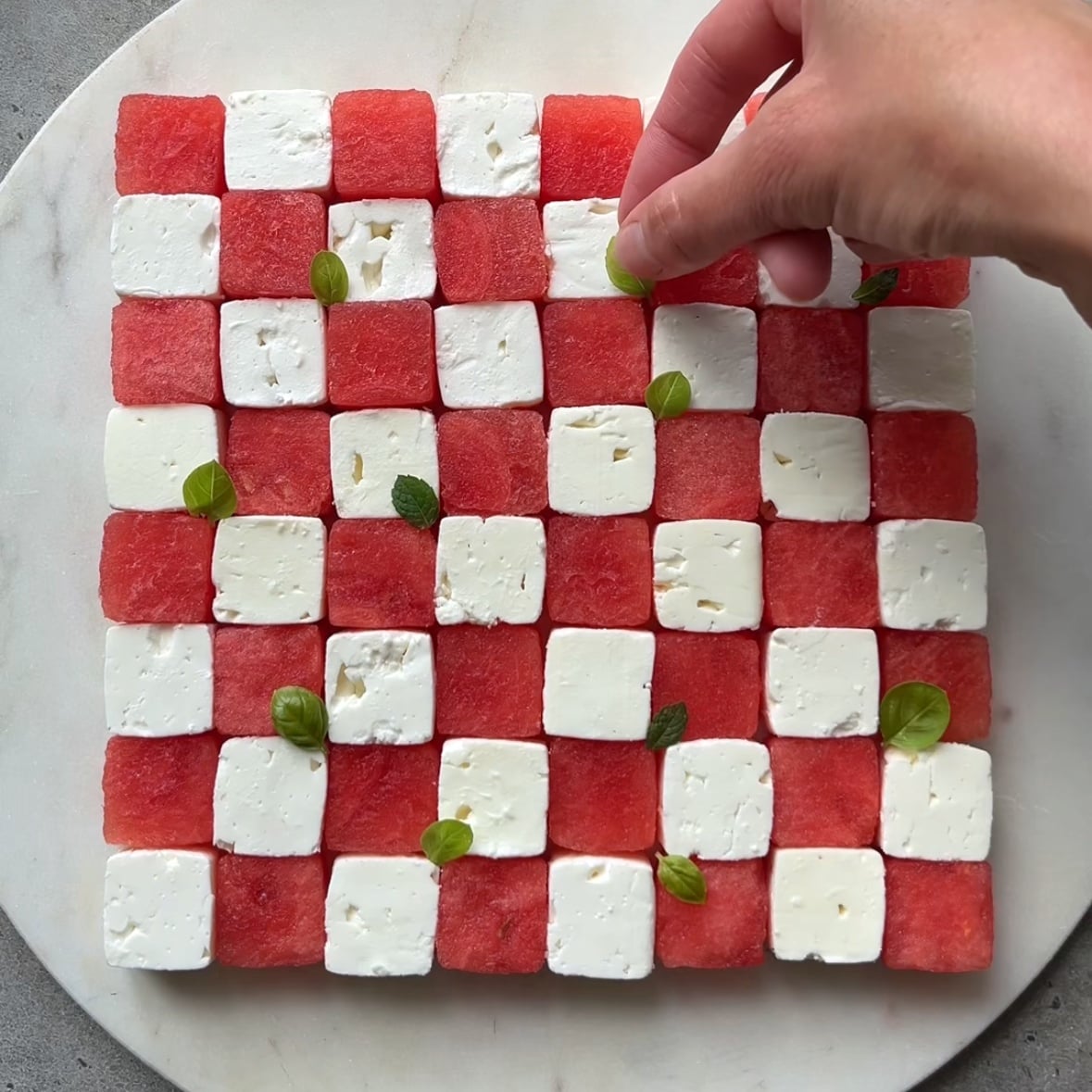 A hand places fresh basil leaves on top of the cubed watermelon and feta salad.