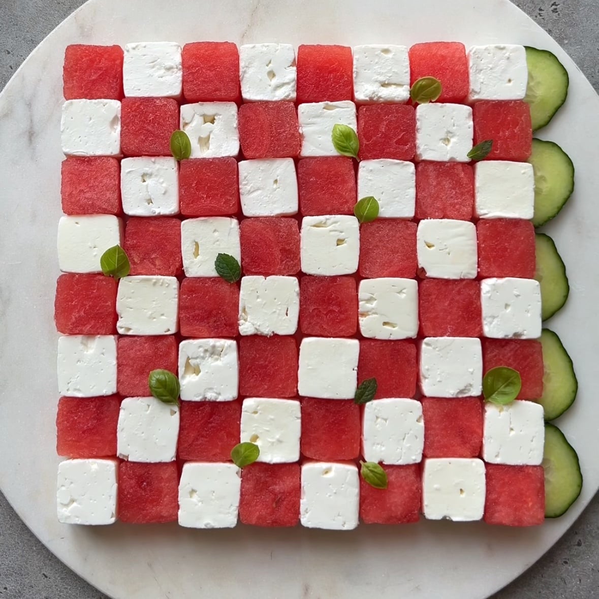 A cubed watermelon and feta salad arranged in a chessboard and topped with fresh basil is being framed with halved cucumber slices.