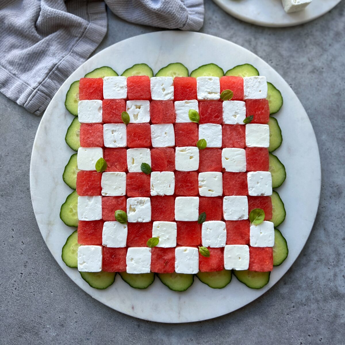 A round marble platter with a checkerboard pattern of watermelon and feta bites, surrounded by cucumber slices and small basil leaves.