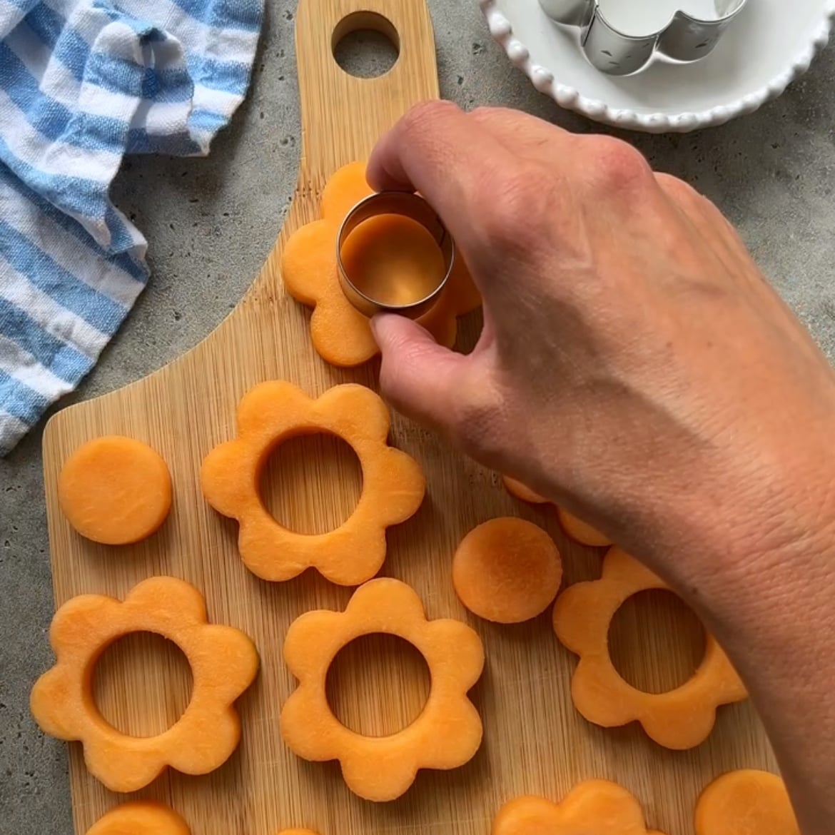 A hand uses a round cookie cutter to cut centers from flower-shaped slices of cantaloupe.