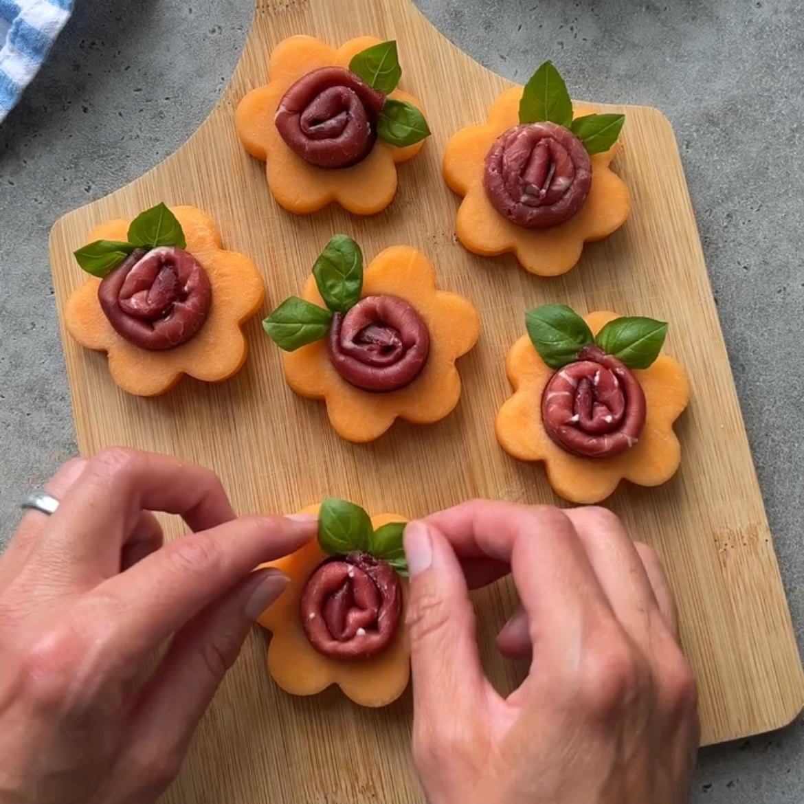 Hands arranging basil leaves on flower-shaped cantaloupe slices topped with rolled cured meat.