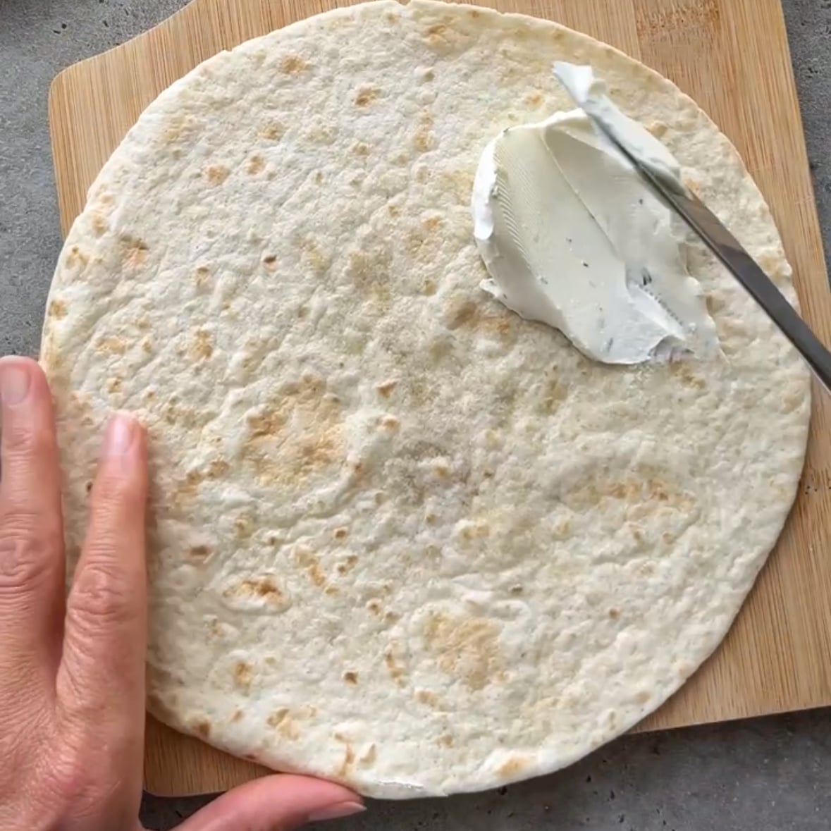 A knife is spreading cream cheese on top of a plain tortilla.