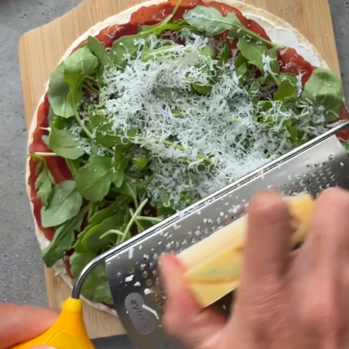 A hand is grating Parmesan on top of a tortilla with cream cheese, bresaola, and arugula.