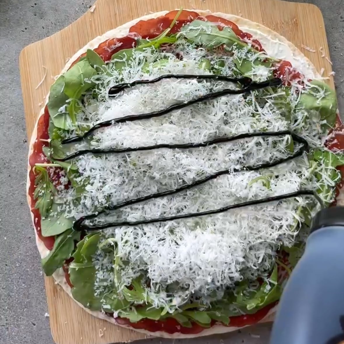 A hand spreads balsamic glaze on top of a tortilla with cream cheese, bresaola, arugula, and finely grated Parmesan.