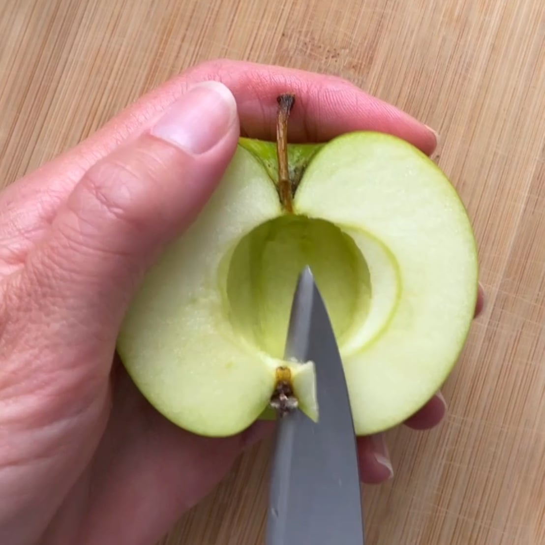 A hand holds a halved green apple while a knife removes the core on a wooden cutting board.