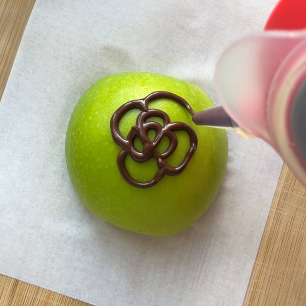 A green apple on parchment paper is being transformed into a Chocolate Covered Apple, with a flower-shaped chocolate design piped on top.
