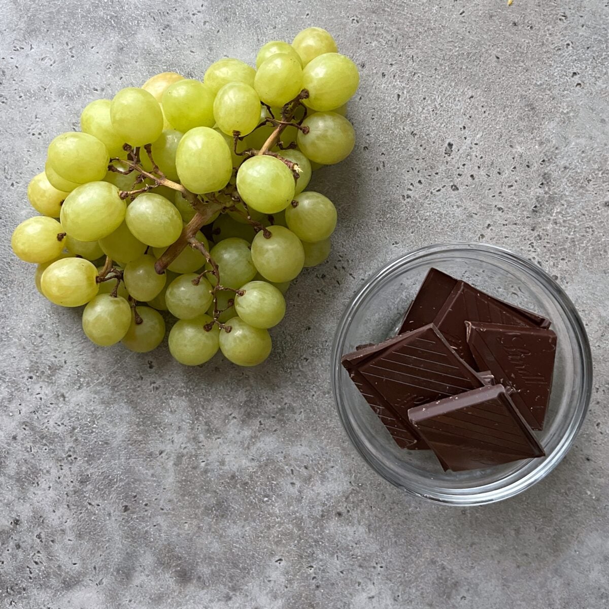 A bunch of green grapes and a small glass bowl with several squares of dark chocolate.