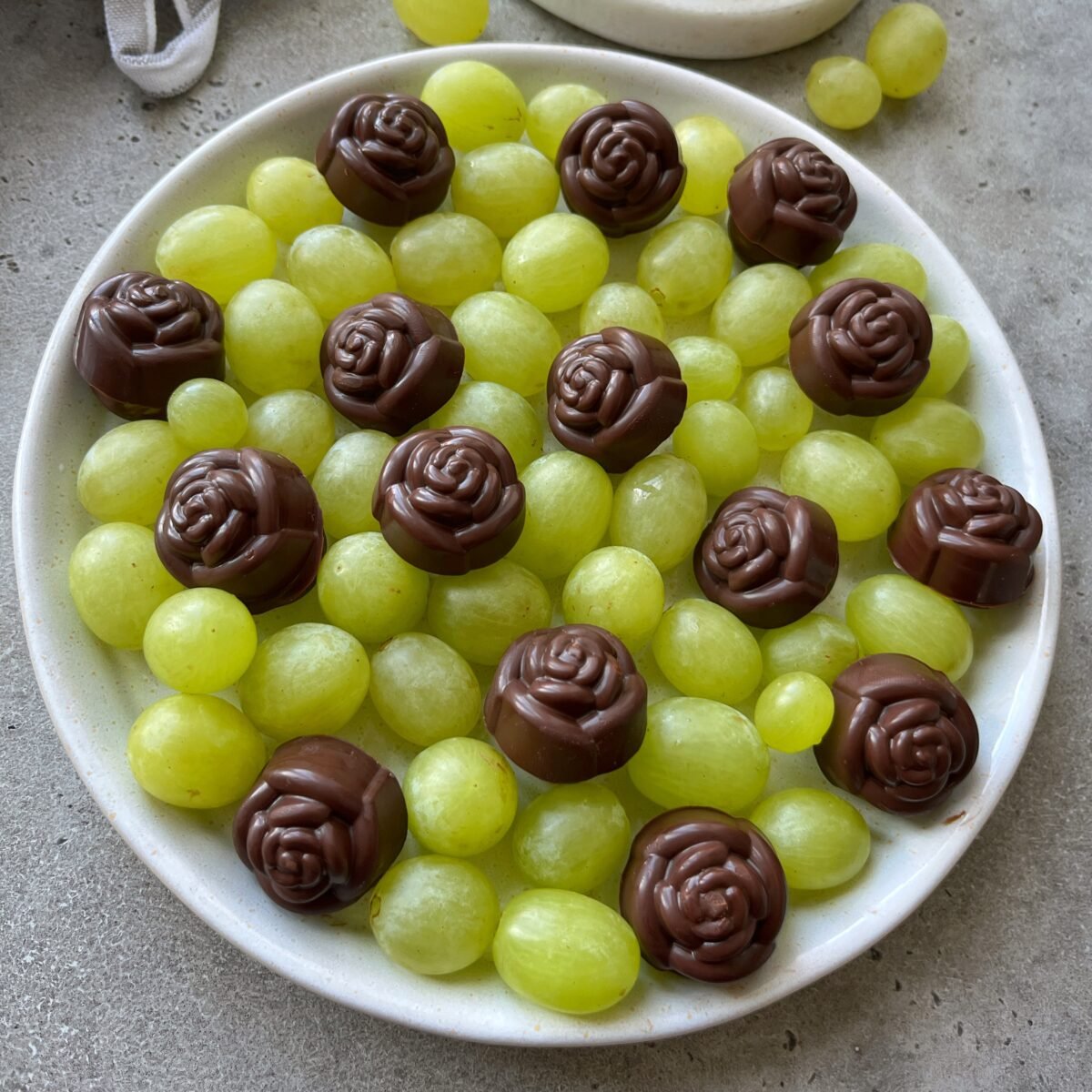 A plate of green grapes topped with several rose-shaped grape chocolates on a gray surface.