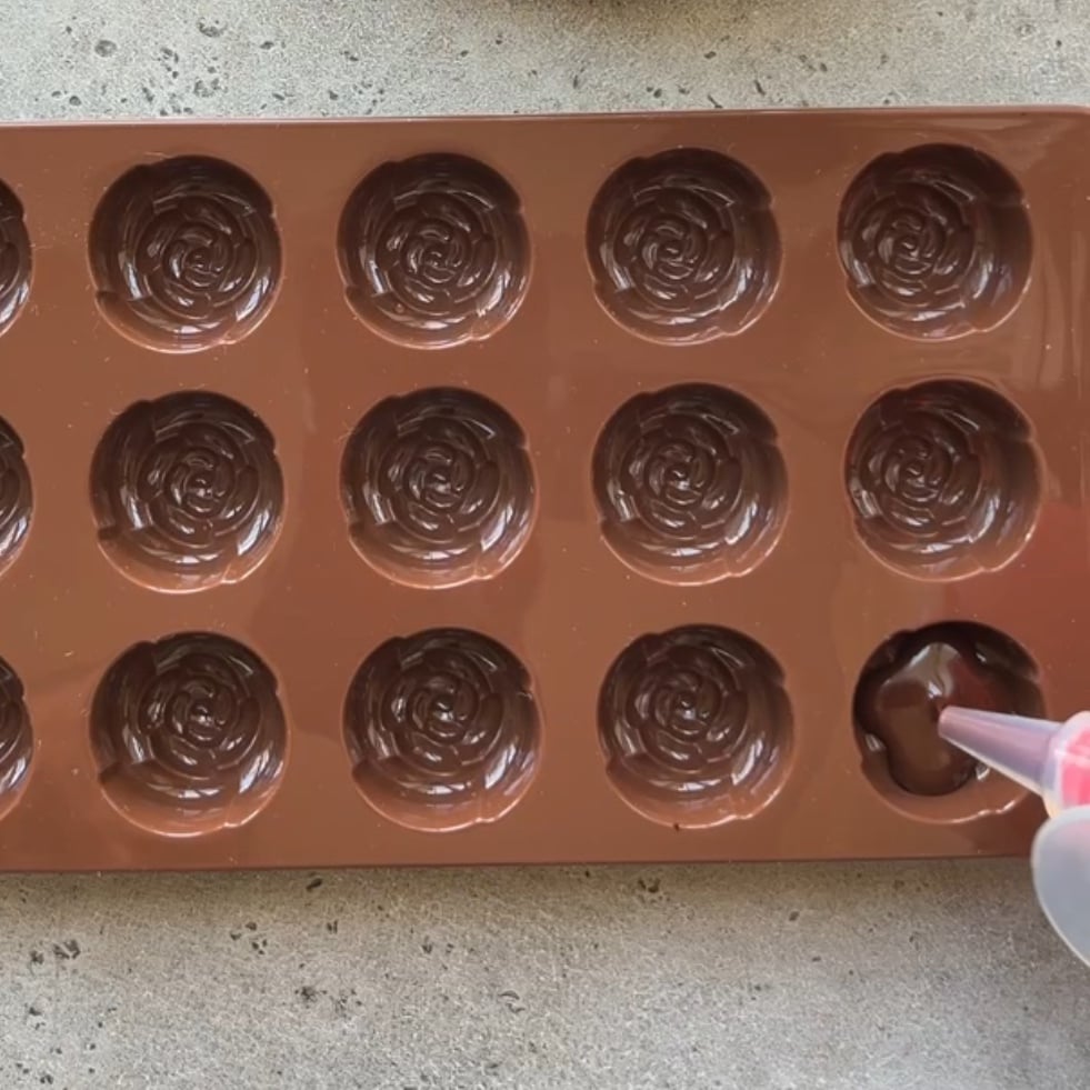 A brown silicone mold with rose-shaped cavities is being filled with chocolate from a piping bag.