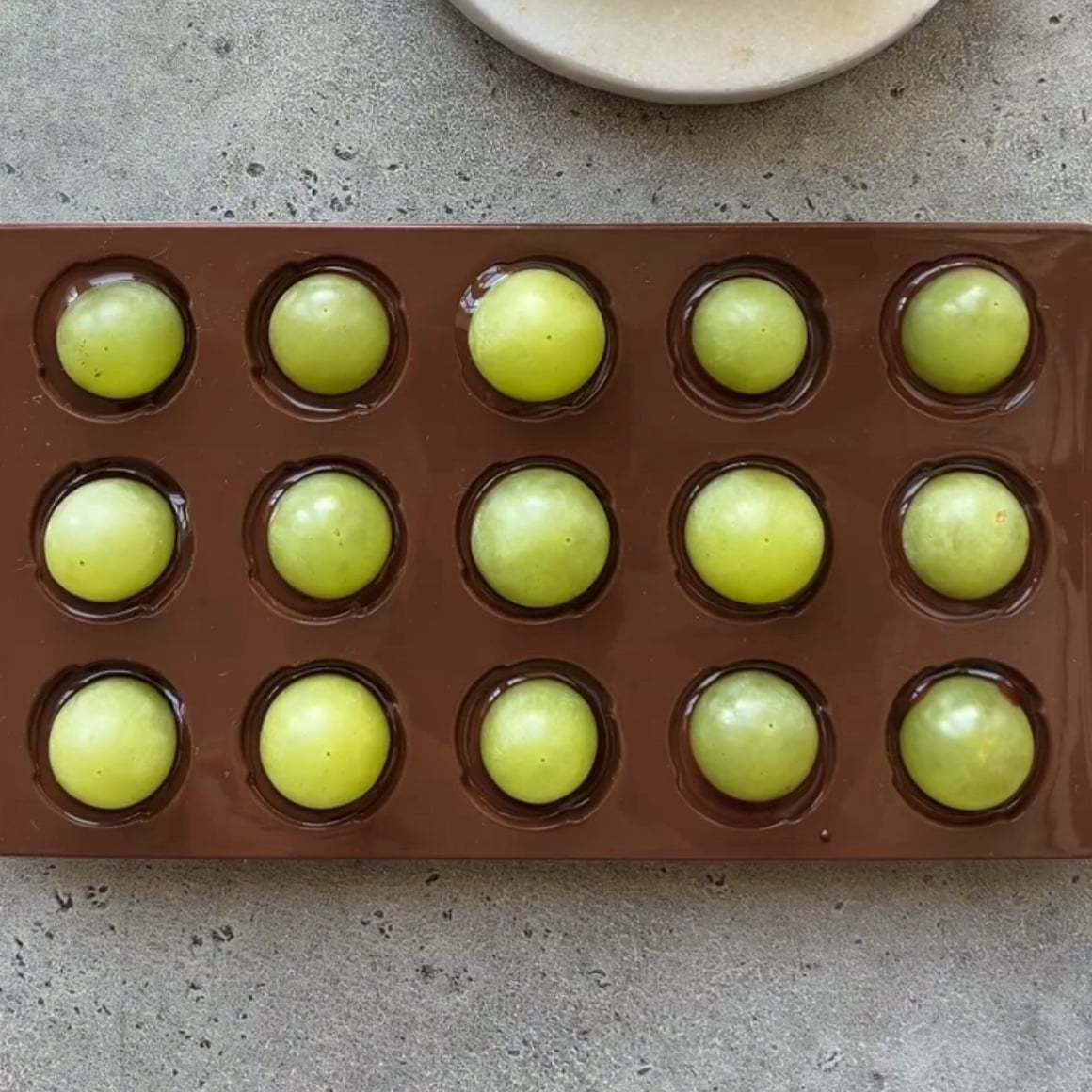 A brown silicone mold is topped with fifteen green grapes in separate circular compartments on a gray surface.