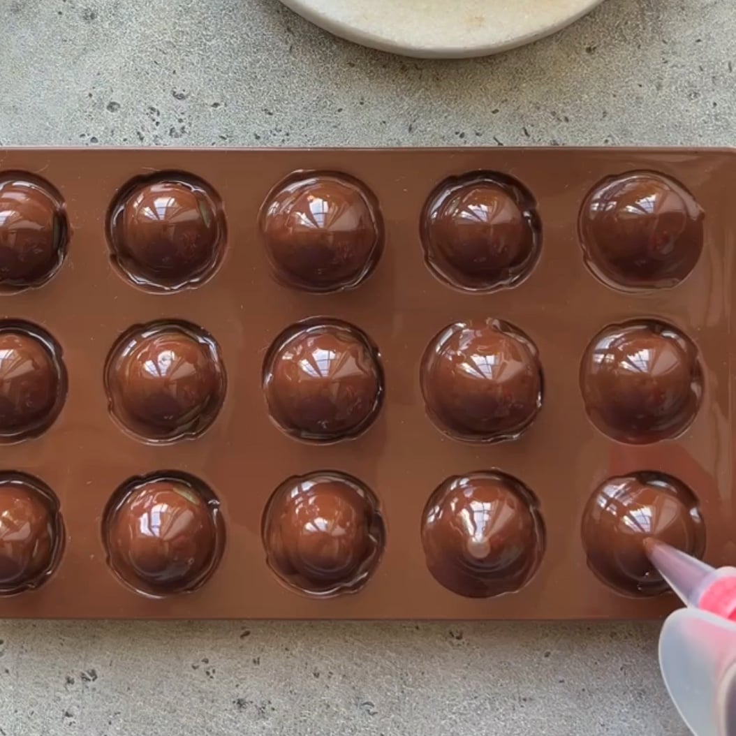 A brown silicone mold holds fifteen glossy, grapes topped with dark chocolate.