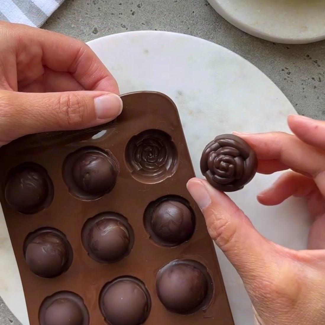 A person holds a grape chocolate molded in a flower shape next to a silicone mold with similar chocolates.
