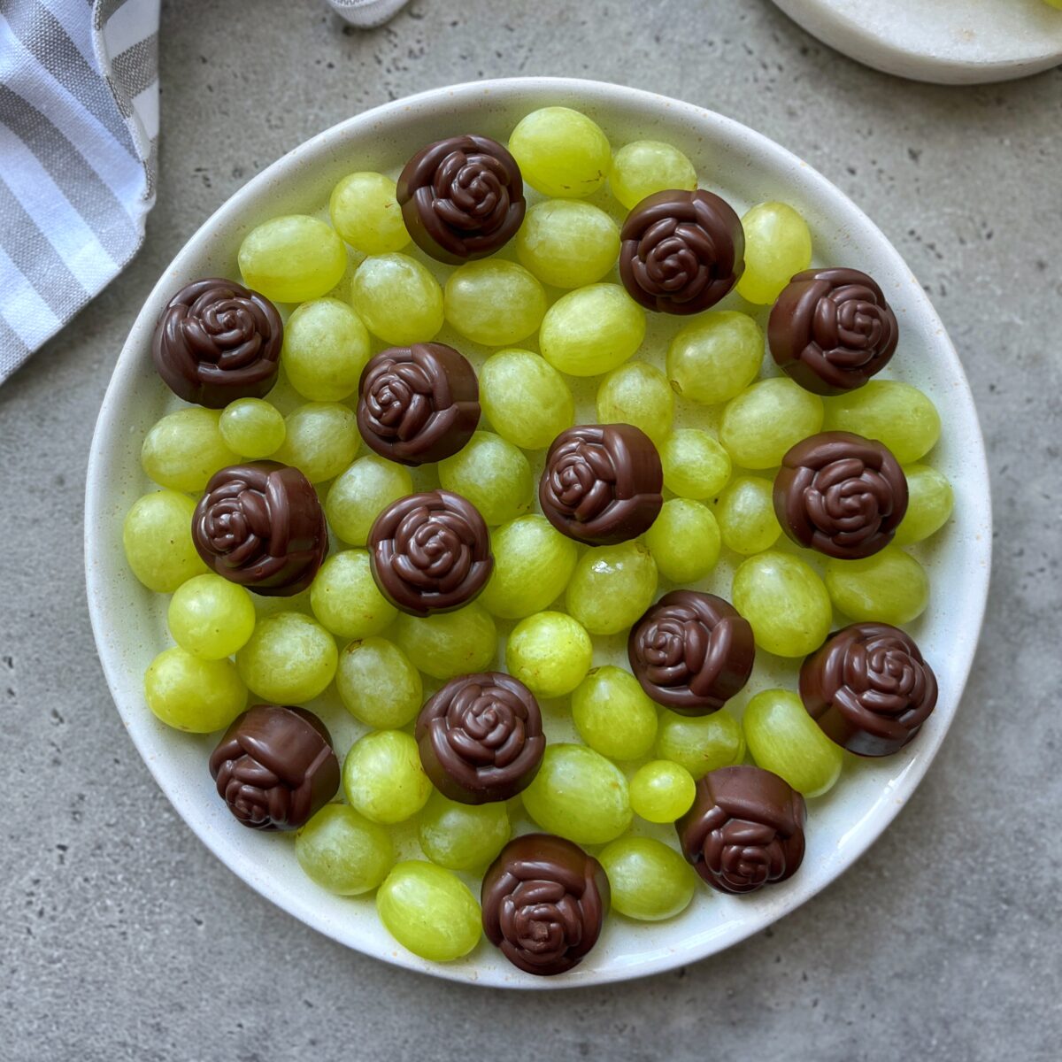 A white plate with green grapes and several rose-shaped chocolate covered grapes arranged on top, set on a gray surface with a striped linen cloth nearby.