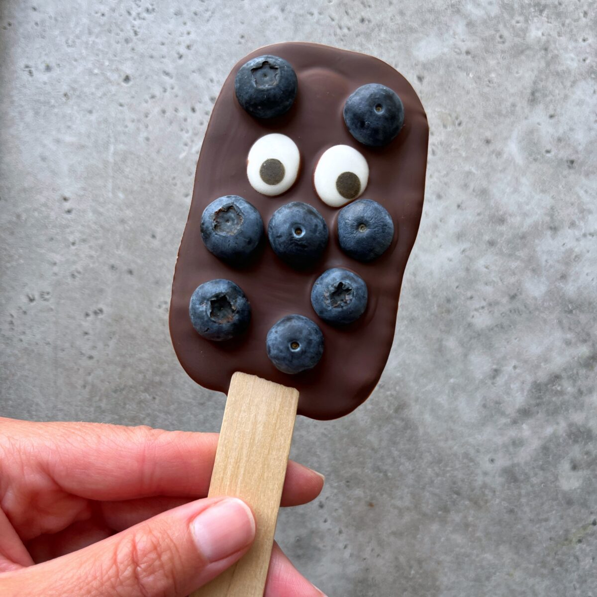 A hand holds a chocolate-covered blueberry lolly topped with two candy eyes on a wooden stick, against a gray surface.
