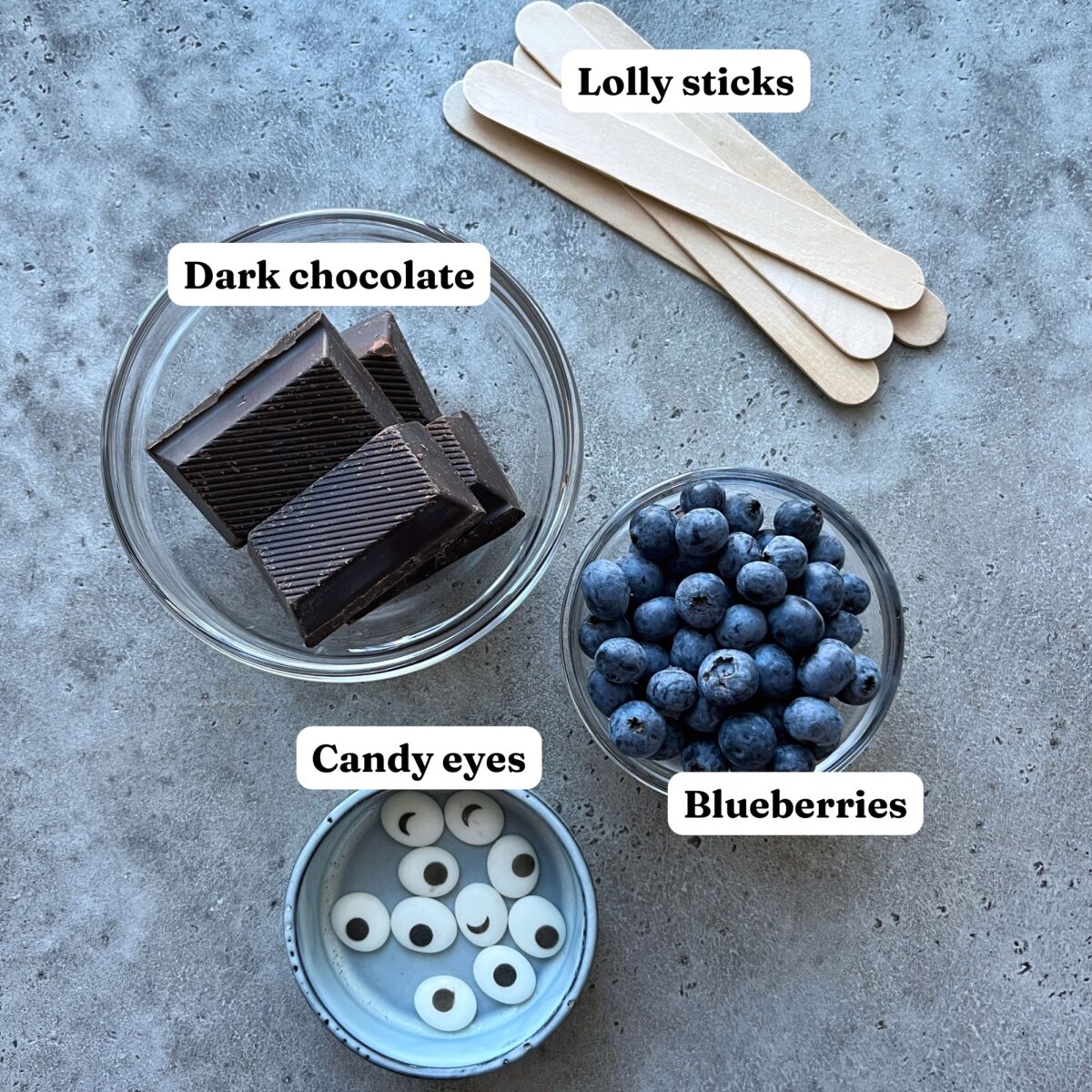 Bowls of dark chocolate, blueberries, and candy eyes, with a stack of wooden lolly sticks.