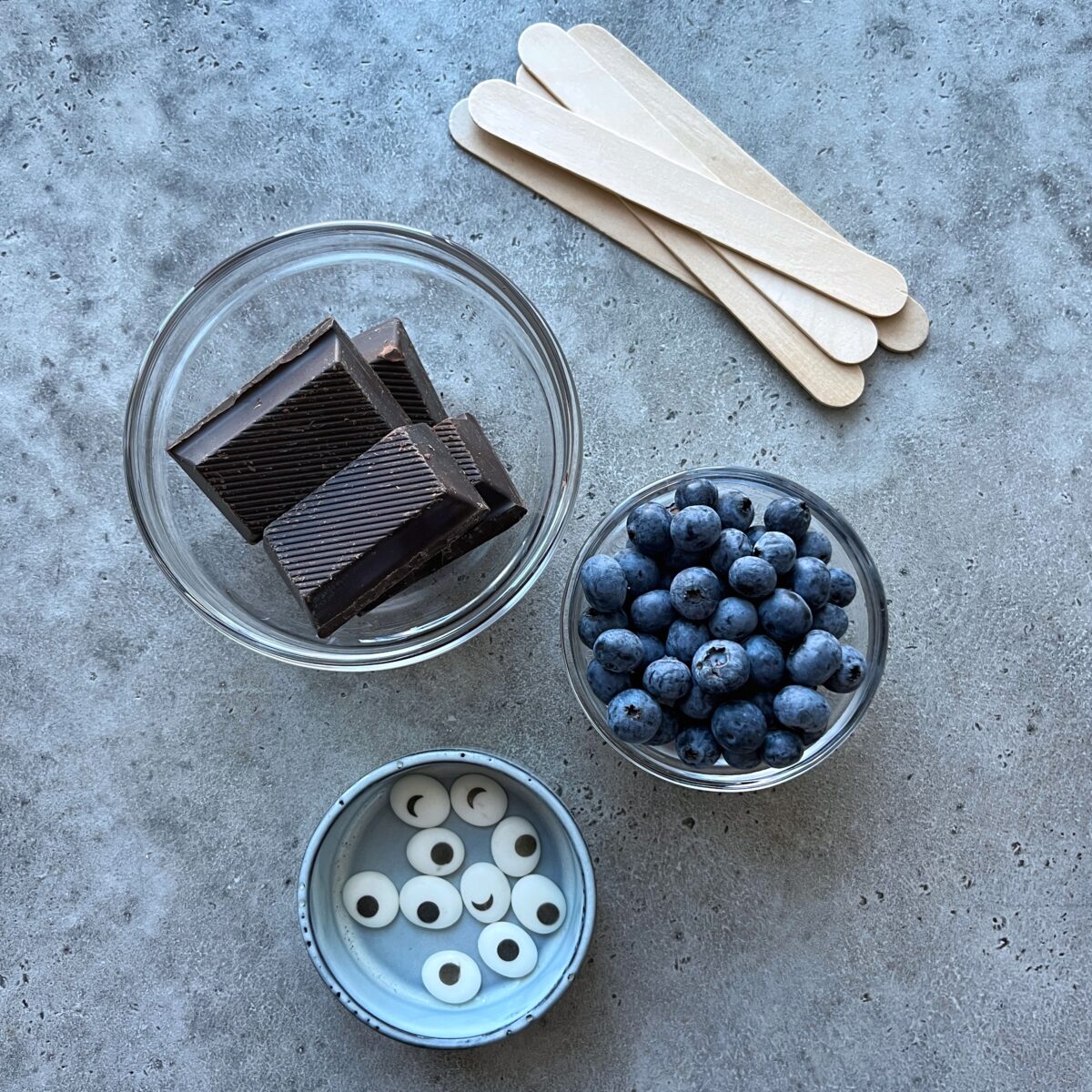 A bowl of chocolate pieces, fresh blueberries, white candy eyes, and wooden sticks on a gray surface.