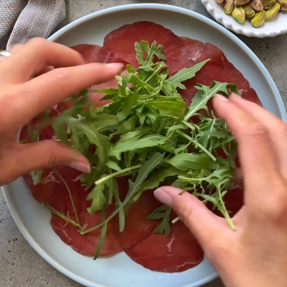 Hands placing fresh arugula on top of thinly sliced cured meat arranged on a round plate, with a small dish of pistachios nearby.