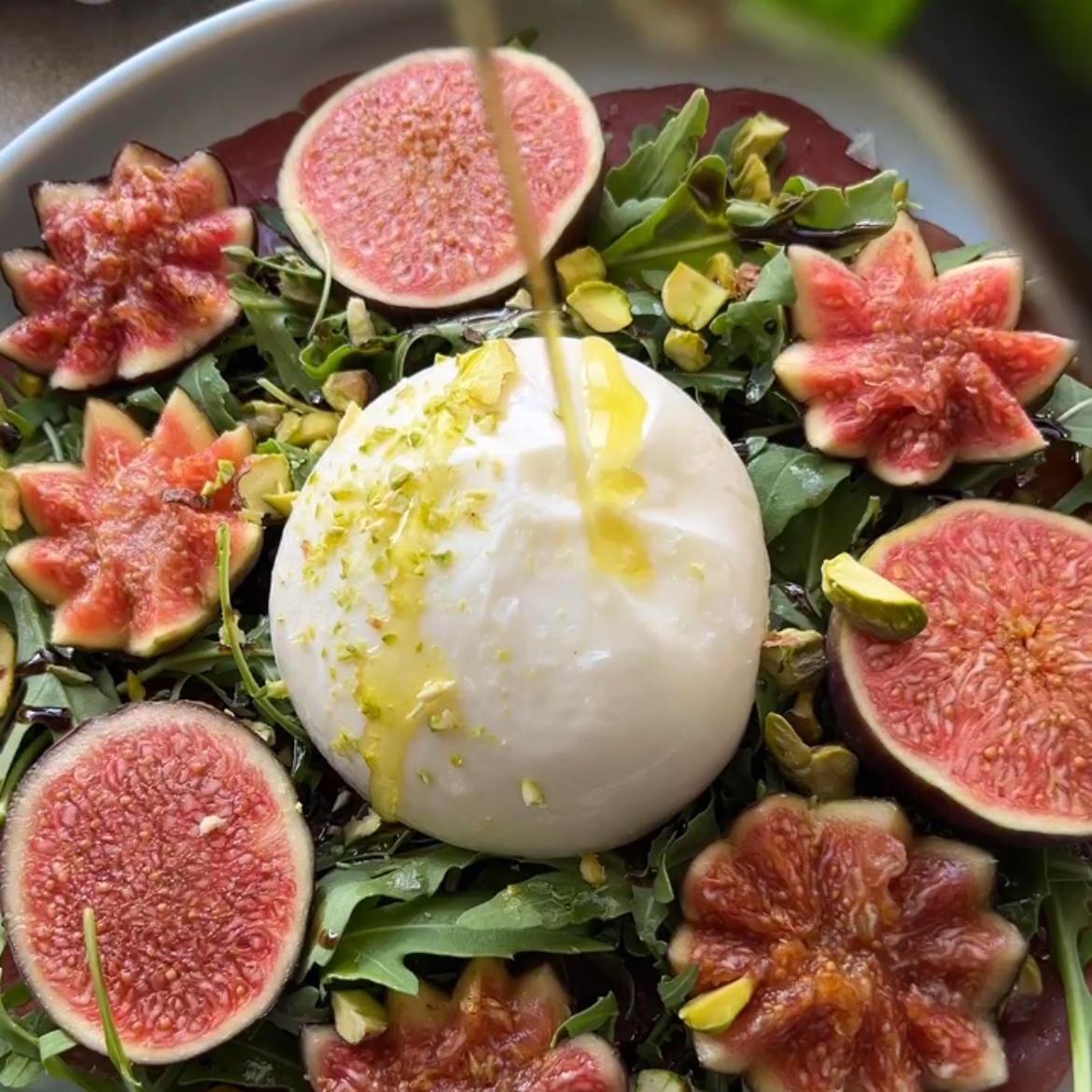Burrata cheese being topped with olive oil on a bed of arugula, surrounded by sliced and decorative-cut figs.
