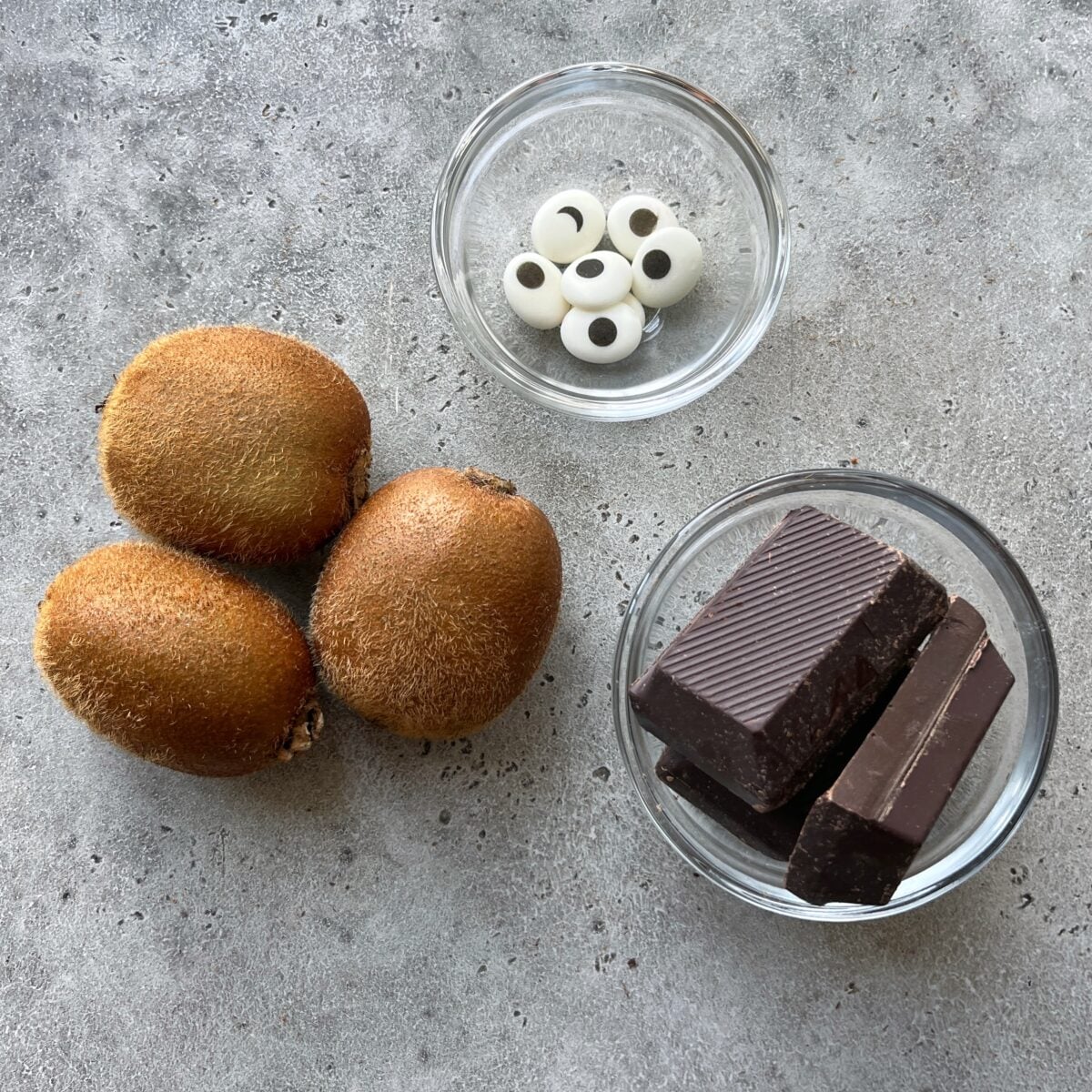 Three whole kiwis, chocolate pieces in a glass bowl, and candy eyes in another bowl are arranged on a gray surface.