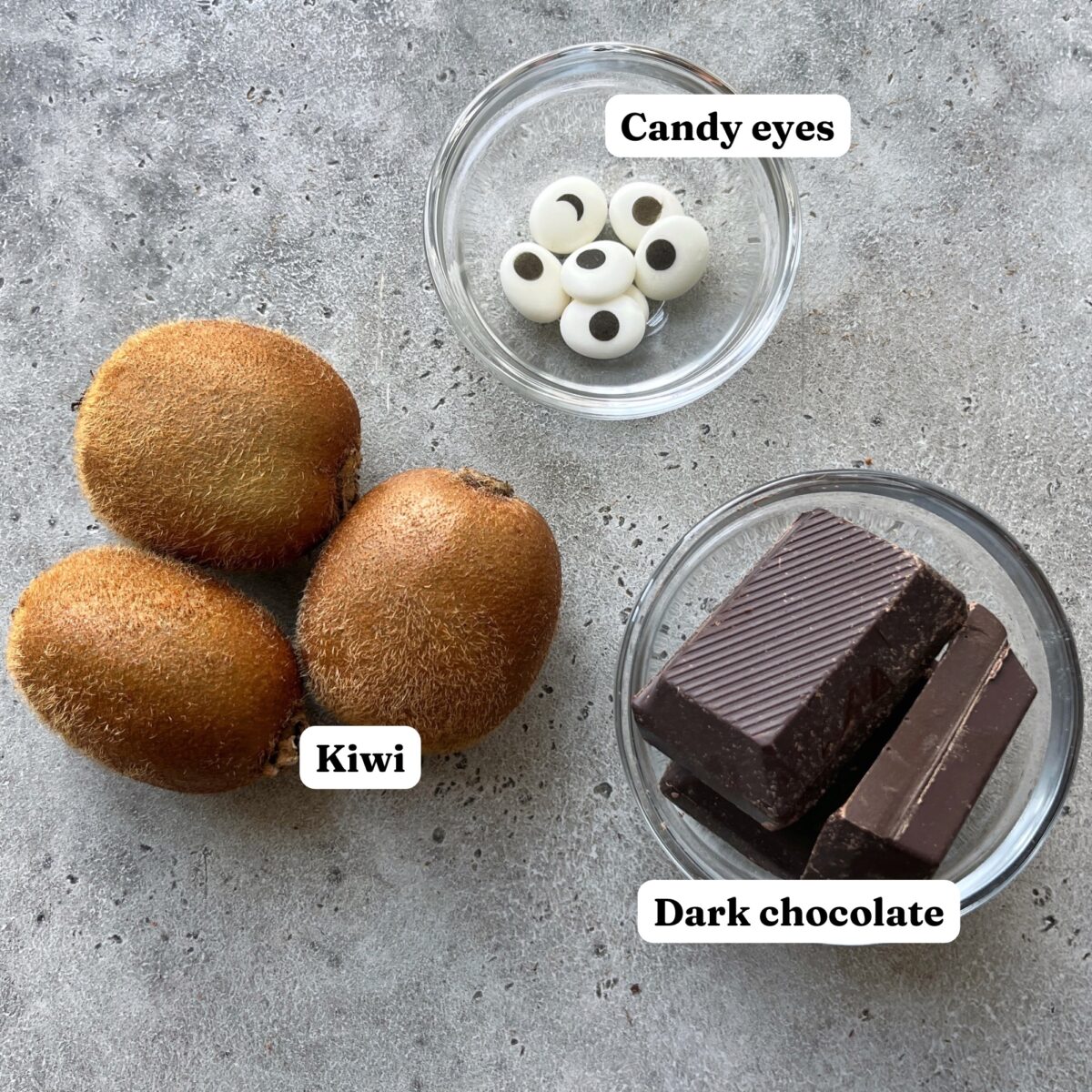 Three kiwis next to small glass bowls containing candy eyes and dark chocolate.