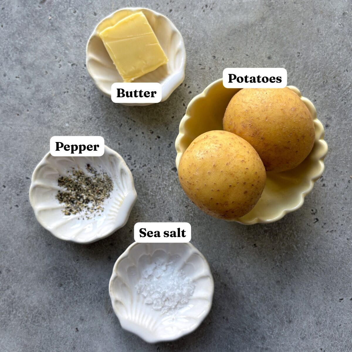 Two whole potatoes, a pat of butter, ground pepper, and sea salt are arranged in small white dishes on a gray surface.
