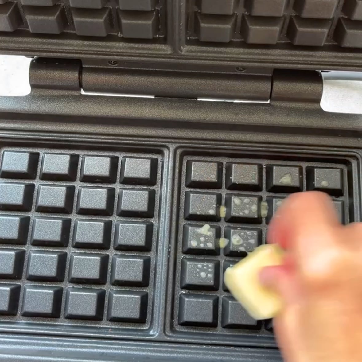 A hand rubs a stick of butter on the griddles of an open waffle maker before use, preparing it for delicious Potato Waffles.