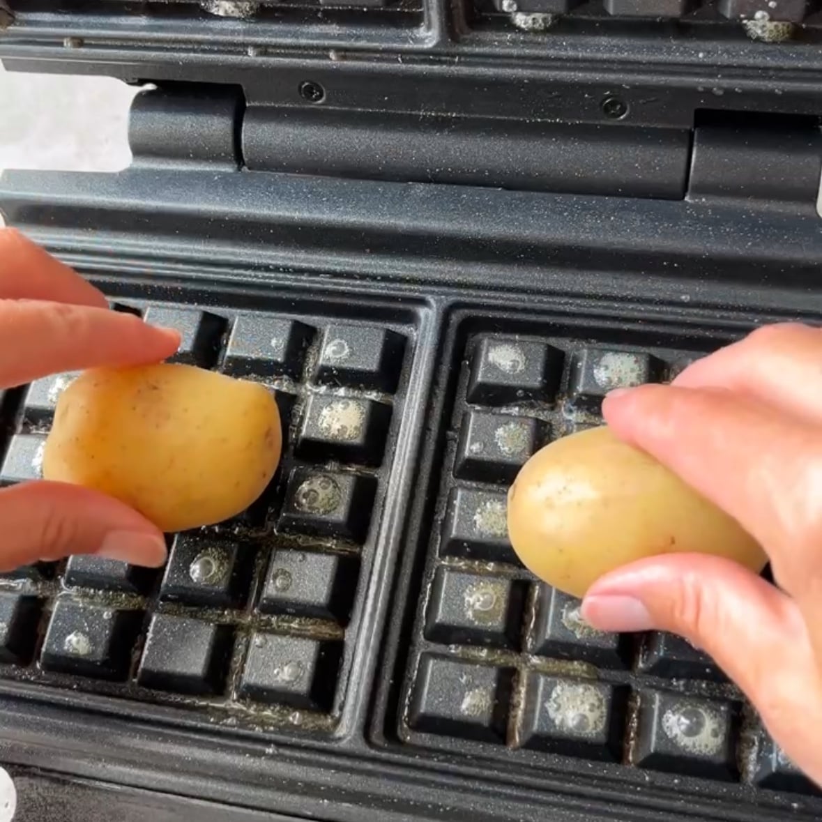 Two hands place small whole potatoes onto the grates of an open waffle maker.