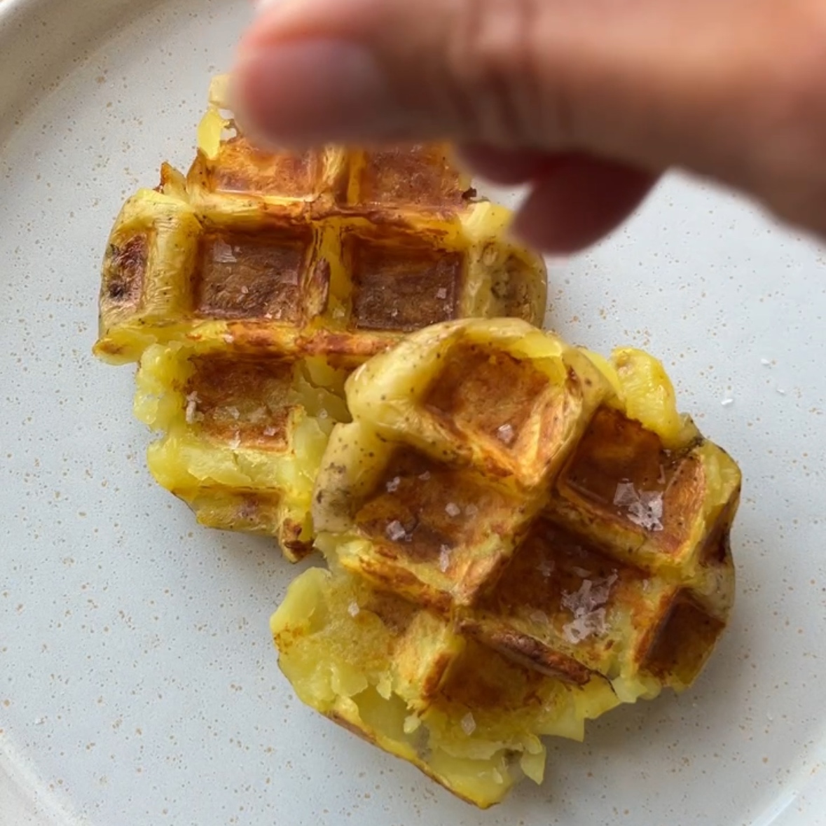 Two crispy Potato Waffles on a plate with a hand sprinkling salt over them.