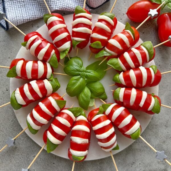 A round marble platter topped with 15 San Marzano tomatoes that are each sliced and filled with bocconcini and basil. Each appetizer is skewered and ready to eat.