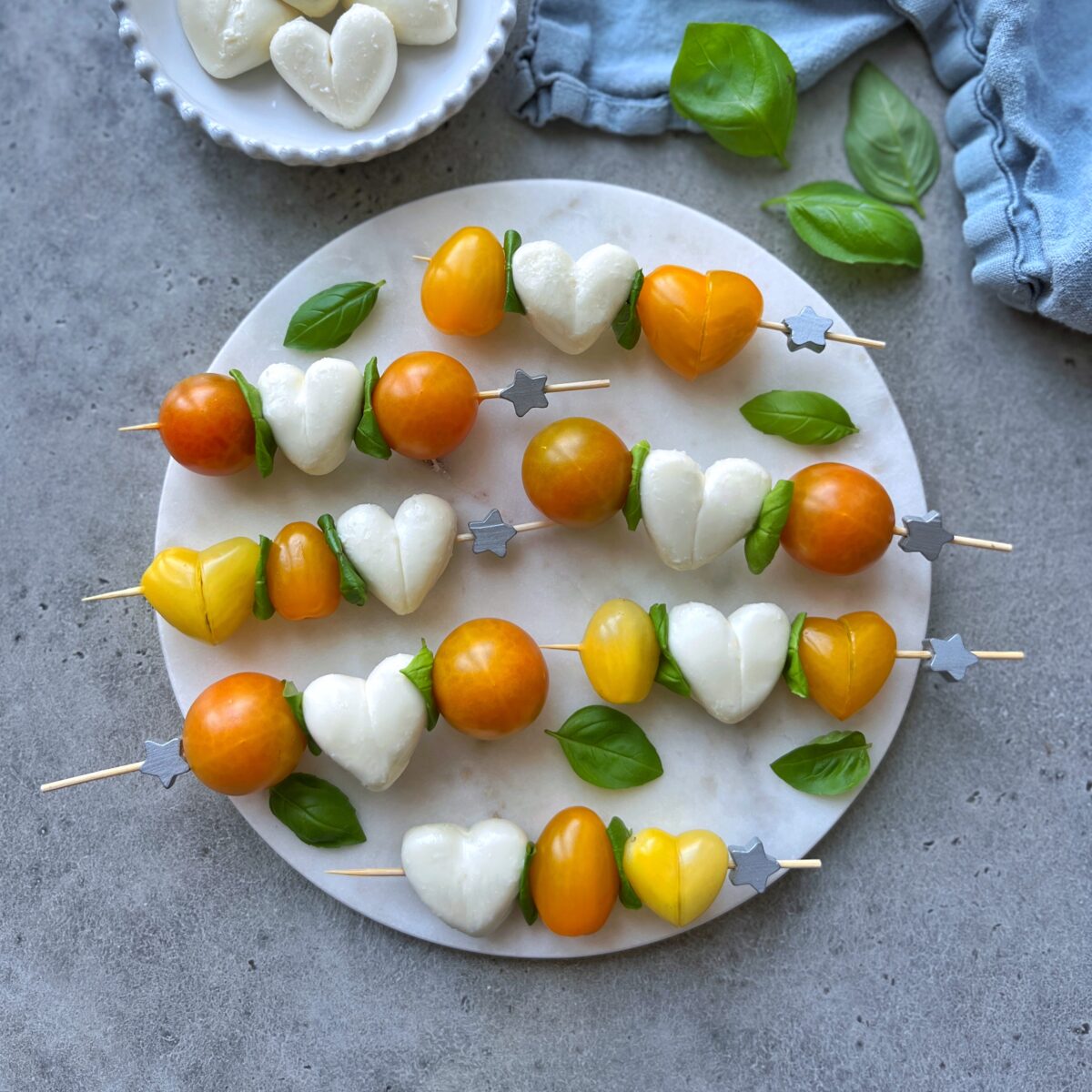 Skewers with heart-shaped mozzarella, yellow and orange cherry tomatoes, and basil leaves with more basil leaves and a blue tea towel nearby.