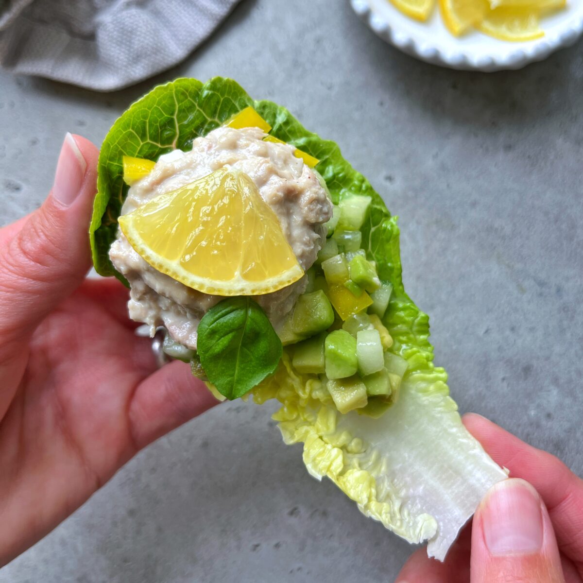 A hand holds one of the tuna salad lettuce boats, topped with diced cucumber, yellow bell pepper, a basil leaf, and a lemon wedge.