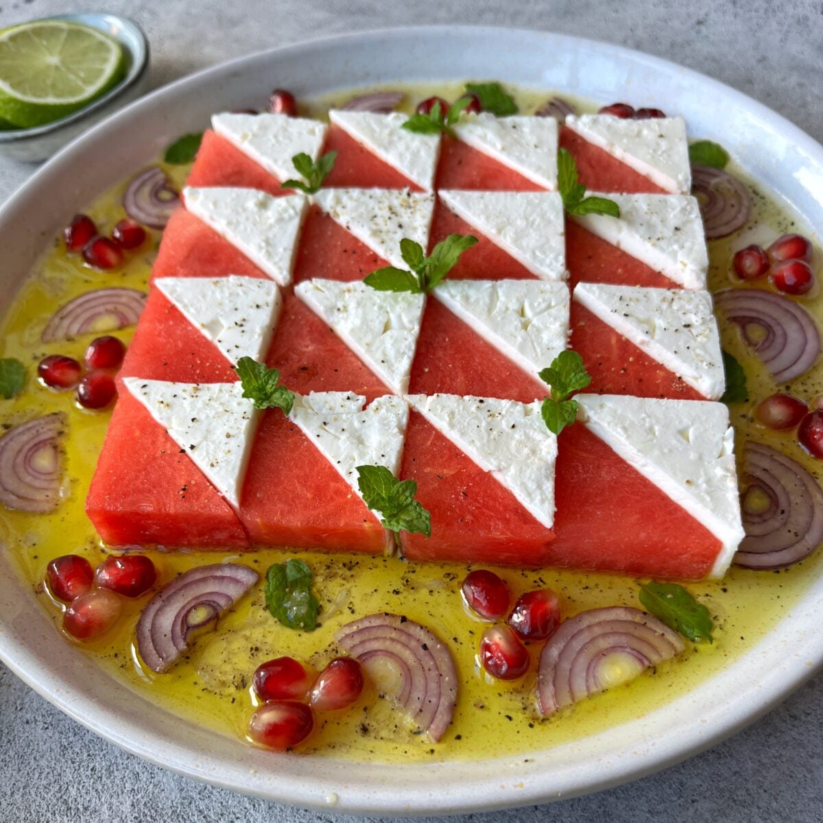 A plate of Watermelon Mint Salad features watermelon and feta triangles arranged in a geometric pattern, garnished with mint, pomegranate seeds, red onion slices, and a drizzle of olive oil lime dressing.