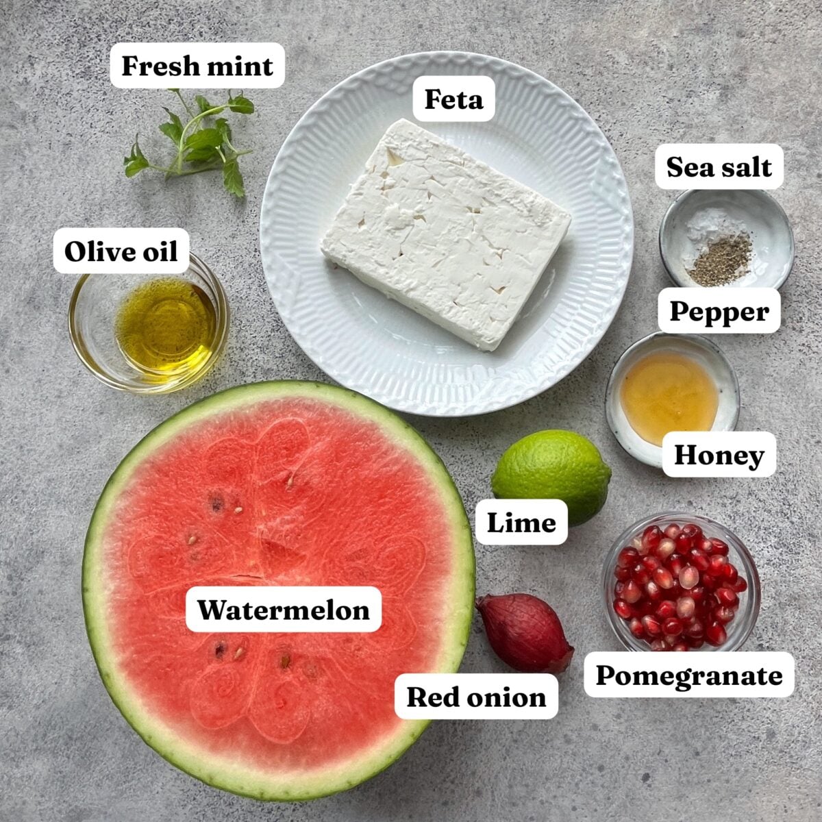 A halved watermelon, feta, fresh mint, sea salt, pepper, olive oil, honey, lime, red onion, and pomegranate seeds are displayed on a gray surface with labels.