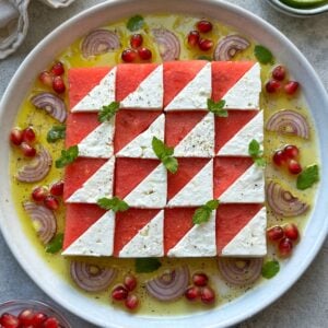 A plate with a geometric Watermelon Mint Salad, featuring alternating triangular pieces of watermelon and feta, garnished with mint, red onion slices, pomegranate seeds, and a citrus dressing.