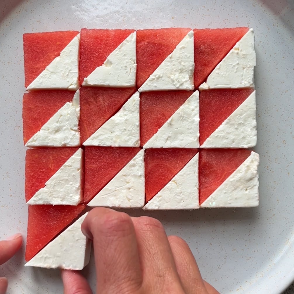 A hand places a triangle of feta cheese and watermelon onto a plate. The pieces are arranged in a geometric grid.