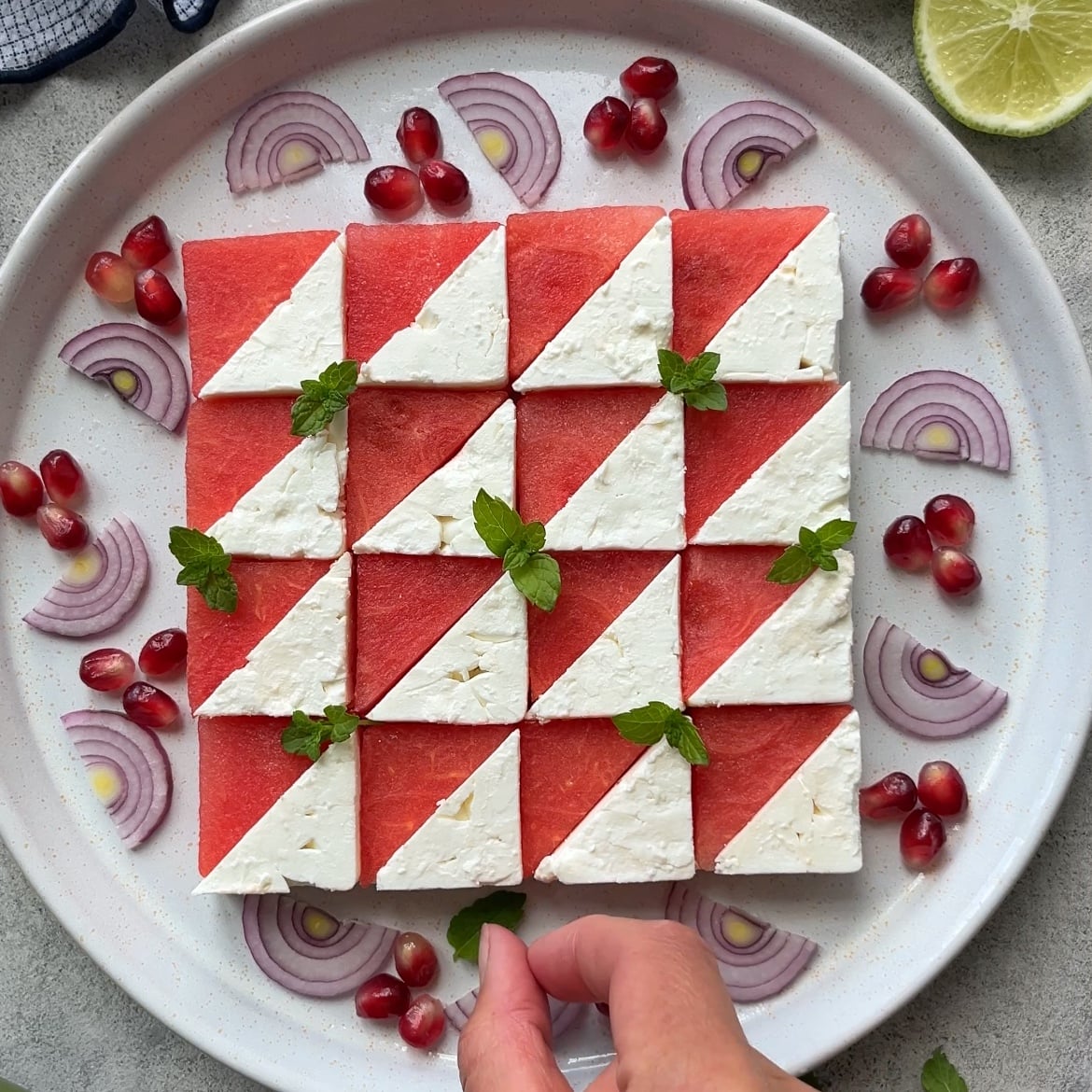 A plate of Watermelon Mint Salad features alternating triangle pieces of watermelon and feta in a checkerboard pattern, garnished with mint, red onion slices, and pomegranate seeds.