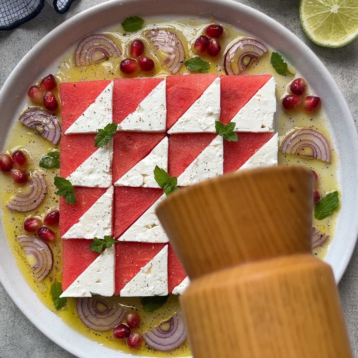 A plate of neatly arranged Watermelon Mint Salad featuring watermelon and feta triangles, red onion slices, pomegranate seeds, mint leaves, and dressing, with a pepper grinder in the foreground.