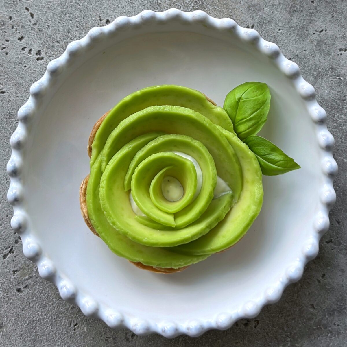 Avocado slices arranged in a rose shape on baked tortilla, garnished with basil leaves, served on a white plate with a textured edge.
