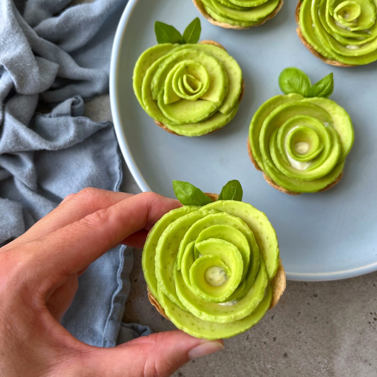 Hand holding an appetizer decorated with avocado slices arranged in a rose shape, with more healthy bites on a plate in the background.