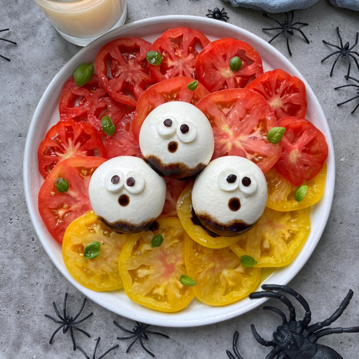 A plate of sliced red and yellow tomatoes topped with three burrata balls decorated as ghost faces, surrounded by plastic spiders.