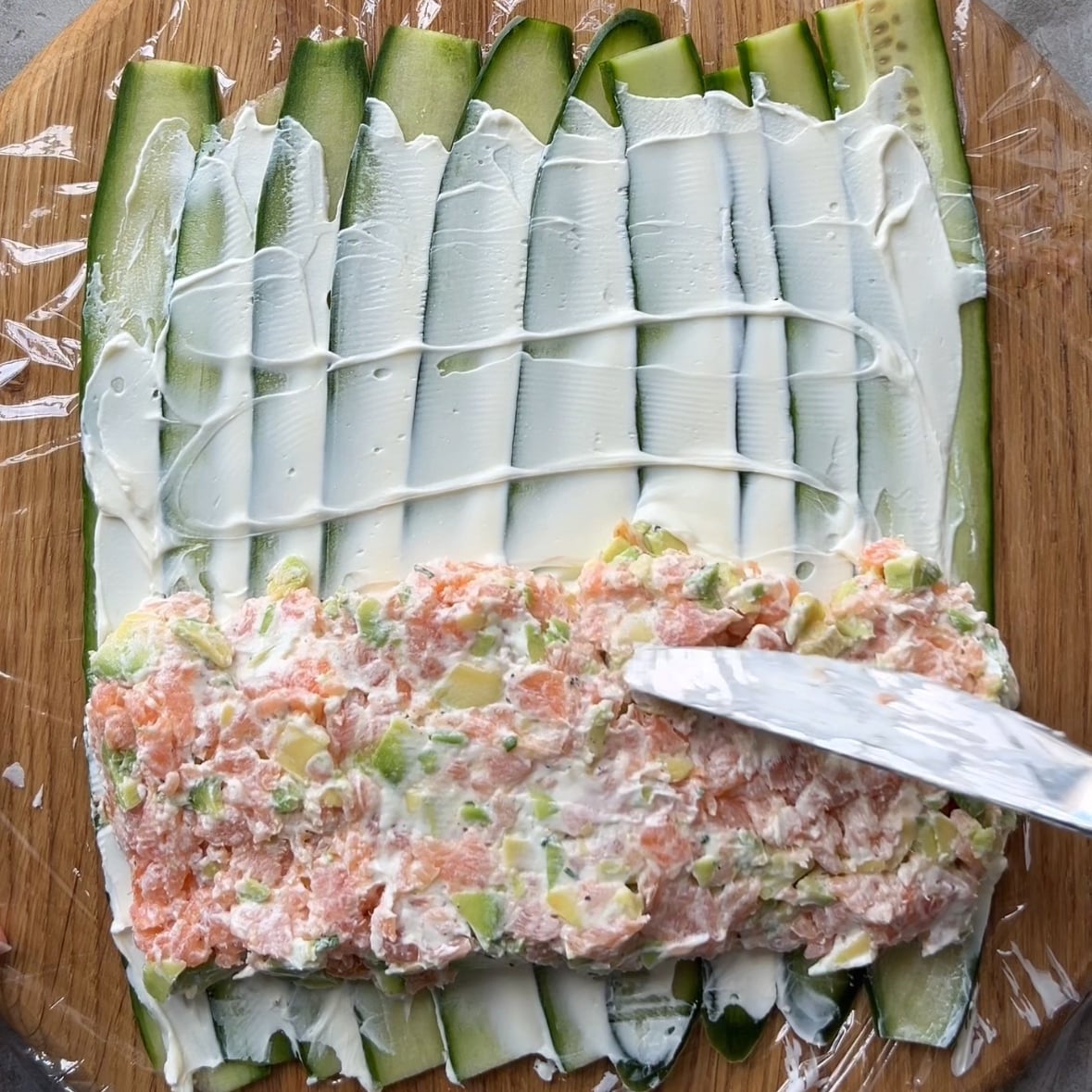 Thin cucumber slices topped with a layer of cream cheese and a savory salmon mixture are being spread with a knife on a wooden board covered with plastic wrap.