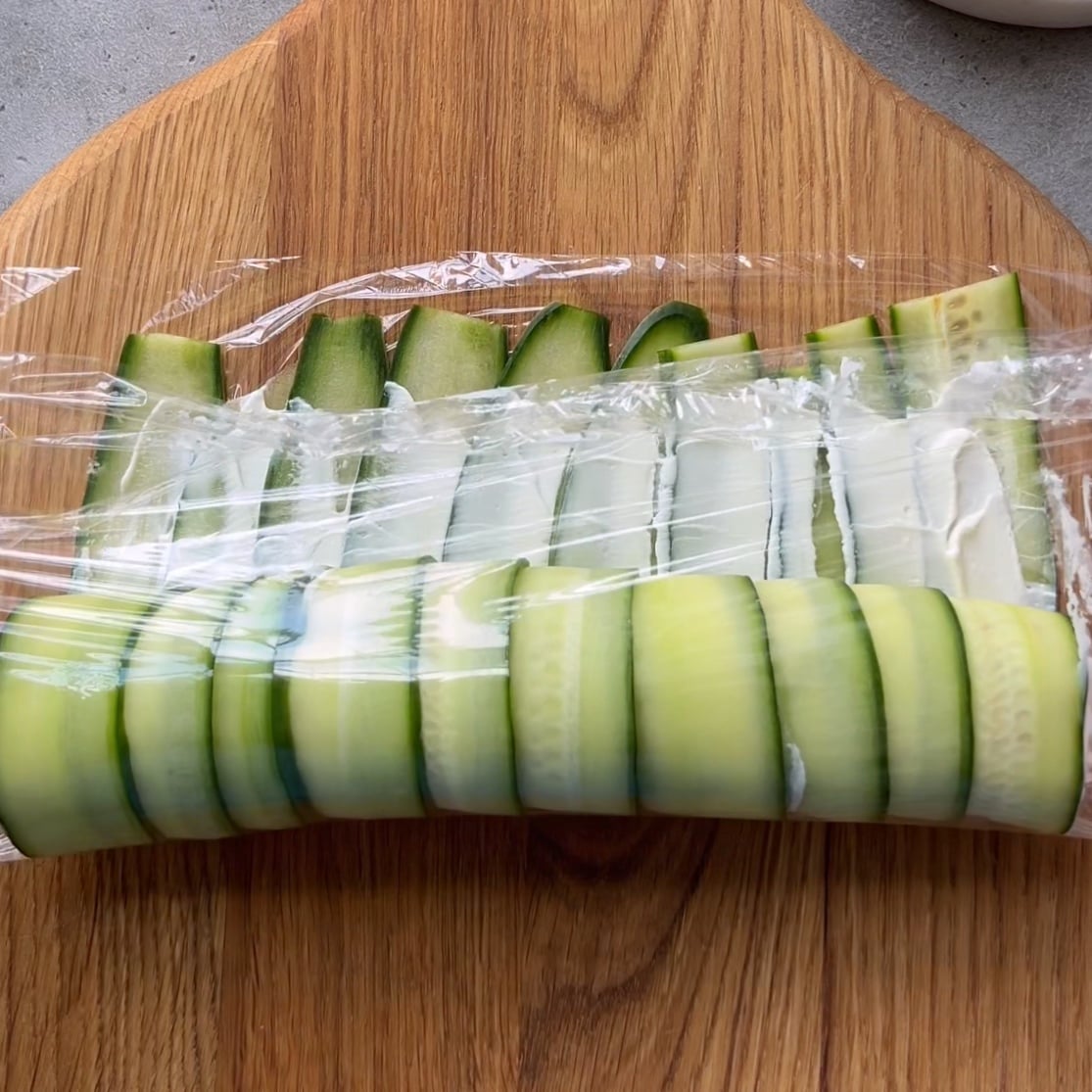 Thin cucumber slices laid out in rows on a wooden board, partially covered with plastic wrap, with a layer of white spread beneath the wrap.